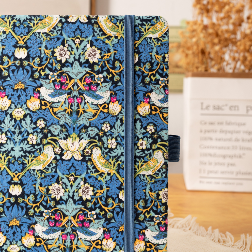 Close-up of the blue bird and floral fabric notebook cover, showcasing the intricate woven bird and botanical pattern, a matching blue elastic closure, and a side pen loop, styled with dried flowers and a kraft paper bag in the background.