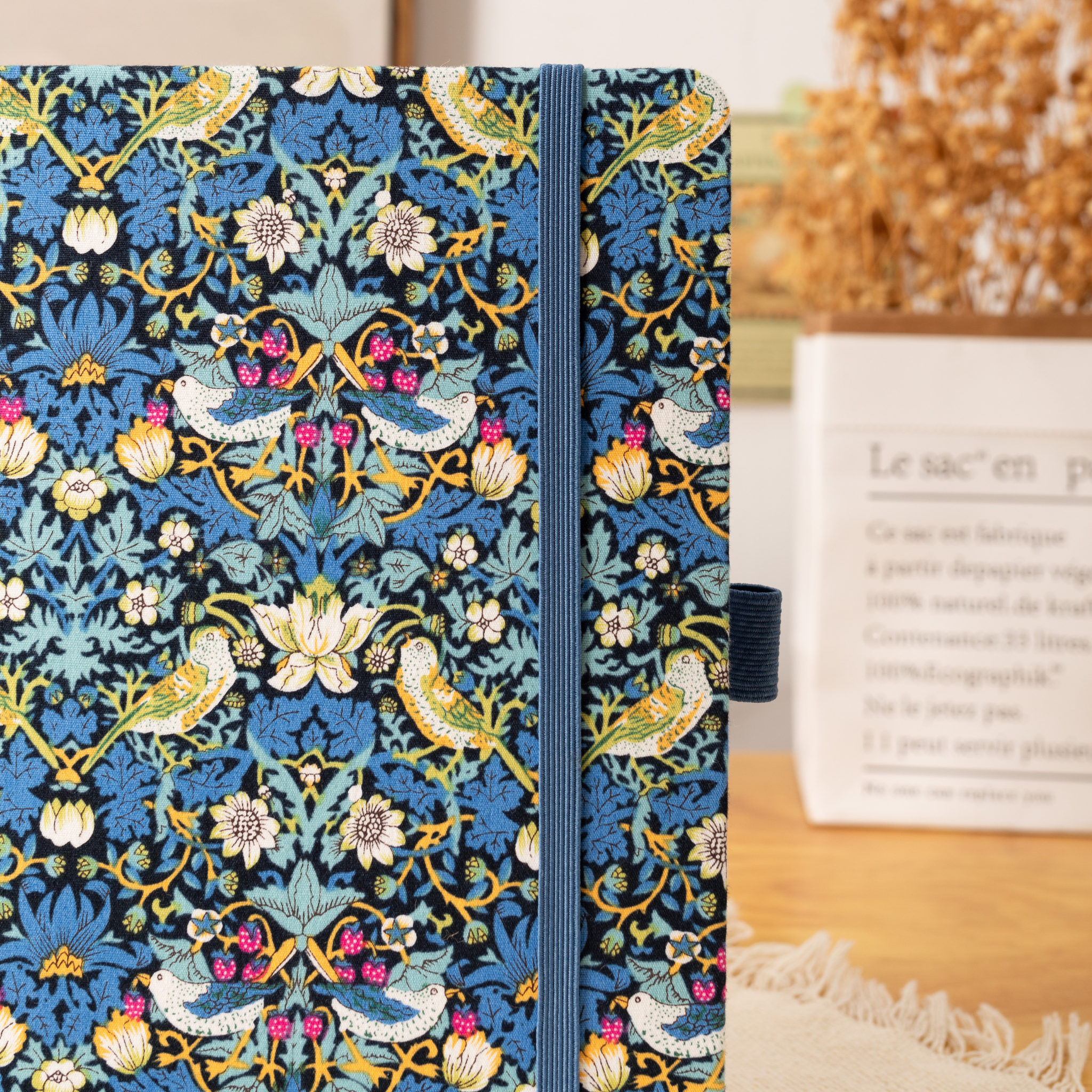 Close-up of the blue bird and floral fabric notebook cover, showcasing the intricate woven bird and botanical pattern, a matching blue elastic closure, and a side pen loop, styled with dried flowers and a kraft paper bag in the background.