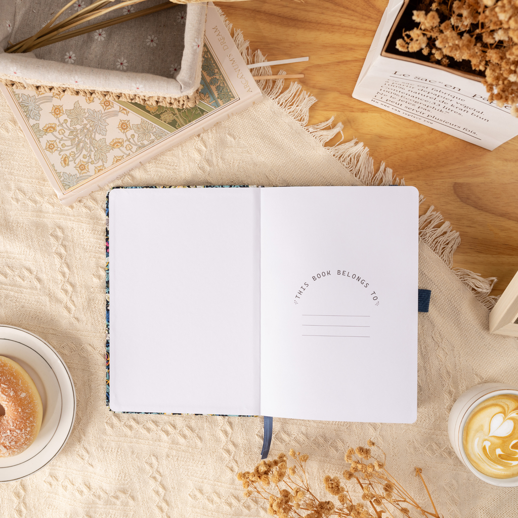 Open blue bird notebook showing the "THIS BOOK BELONGS TO" ownership page, laid flat on a cream surface with a matching ribbon bookmark.