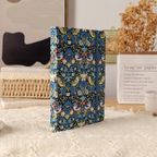 Blue bird pattern fabric notebook standing upright on a cream textured blanket, styled with a book and dried grass.