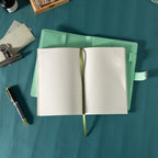 Top-down view of the open green vegan leather journal lying flat on a dark teal surface, showing the lined paper interior with a green ribbon bookmark, an open metal lock strap, and styled with a patterned pen, stamps, and ink.