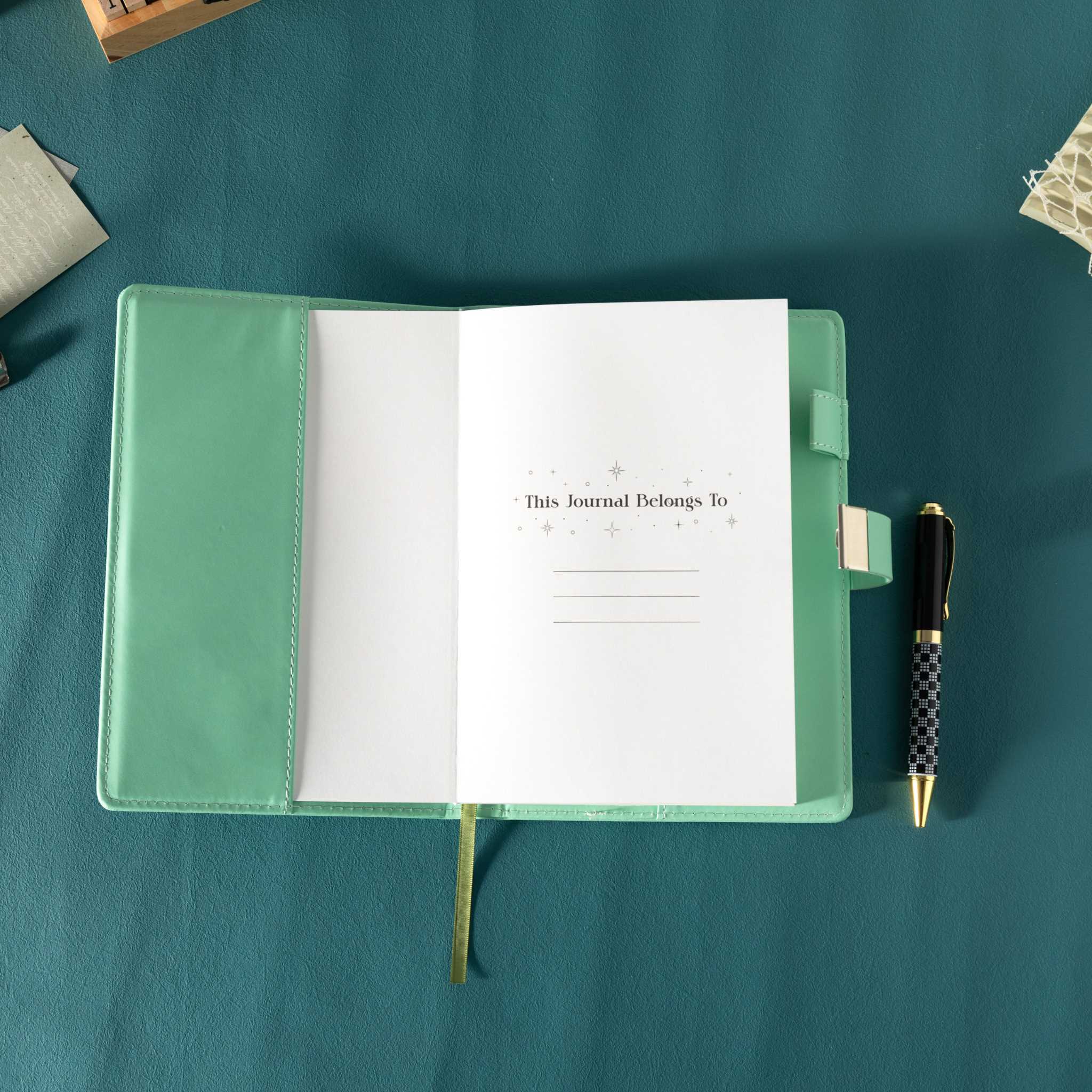 Open green vegan leather notebook showing the "This Journal Belongs To" ownership page, laid flat with a green ribbon and a patterned pen.