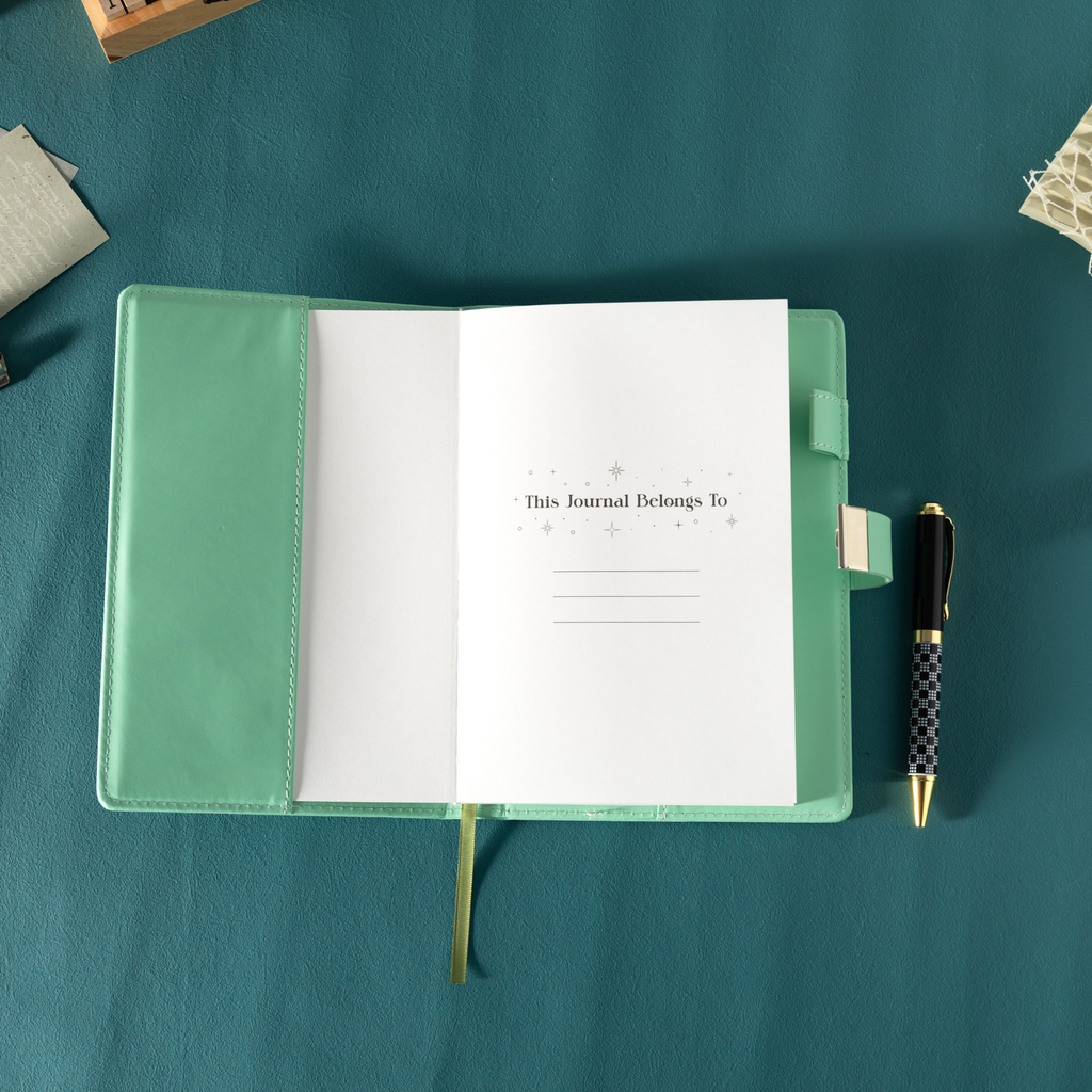 Open green vegan leather notebook showing the "This Journal Belongs To" ownership page, laid flat with a green ribbon and a patterned pen.