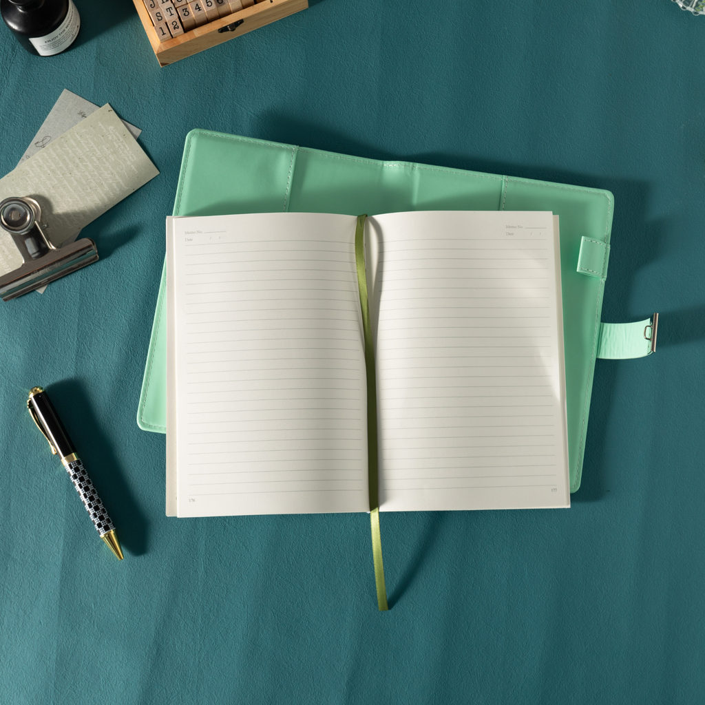 Top-down view of the open green vegan leather notebook lying flat on a dark teal surface, showing the lined paper interior with a green ribbon bookmark, an open metal lock strap, and styled with a patterned pen, stamps, and ink.