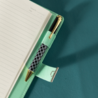 Close-up of the open green vegan leather notebook, showing a black and white patterned pen tucked into the side pen loop, with the metal lock strap extended, and lined paper with a page number visible.