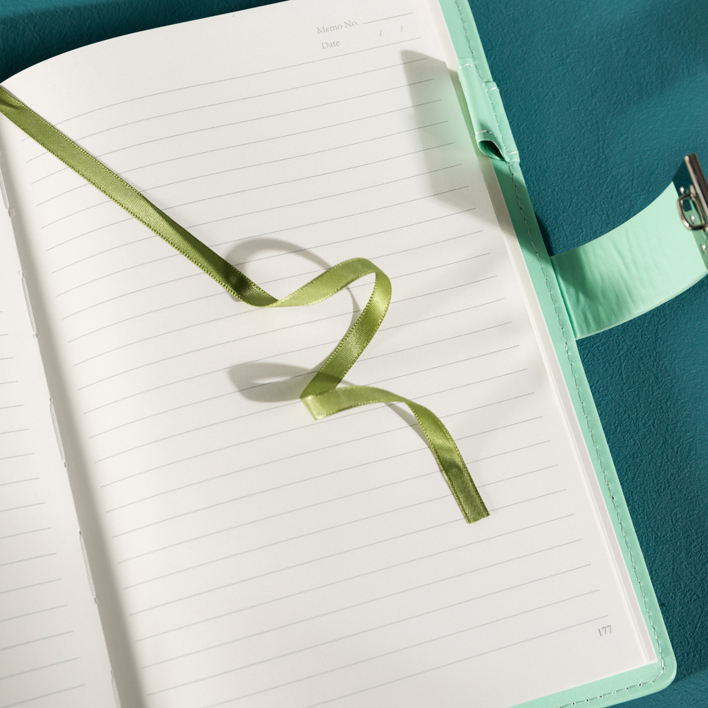 Close-up of the open lined paper interior with a green satin ribbon bookmark and a patterned pen.