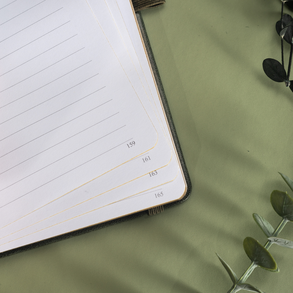 Close-up of the inner page corner of the dark green vegan leather notebook, showcasing layered lined paper with gold borders, corner page numbers, and a dark green leather edge, styled with botanical shadows.