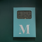 Light blue window-cut gift box featuring a white "M" logo, displaying the dark green mountain vegan leather notebook inside.