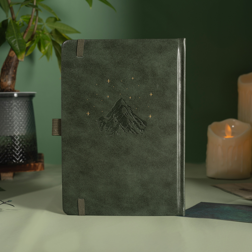 Back cover of the dark green vegan leather notebook standing upright, featuring a subtle embossed mountain design with small gold foil stars, a side pen loop, and matching green elastic band, styled with a potted plant and candle. 