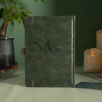 Back cover of the dark green vegan leather notebook standing upright, featuring a subtle embossed mountain design with small gold foil stars, a side pen loop, and matching green elastic band, styled with a potted plant and candle. 