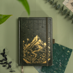 Dark green vegan leather notebook lying flat on a light green desk, showcasing the gold foil mountain and starry night cover design, matching green elastic band, and botanical leaves surrounding it.