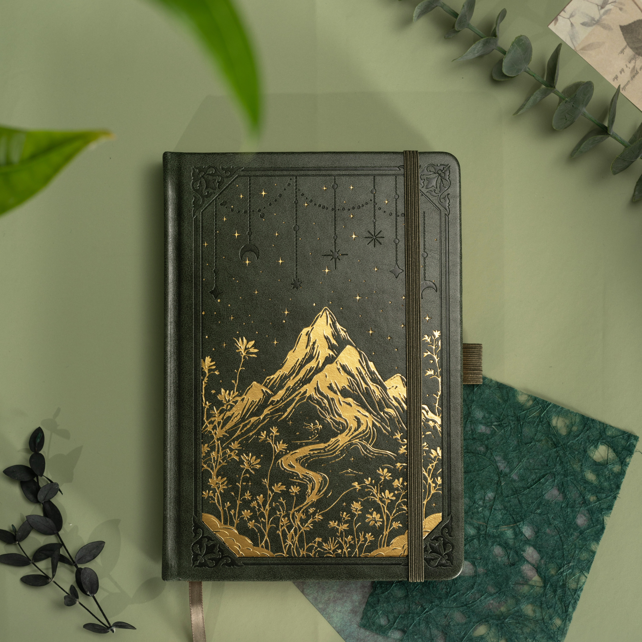 Dark green vegan leather notebook lying flat on a light green desk, showcasing the gold foil mountain and starry night cover design, matching green elastic band, and botanical leaves surrounding it.