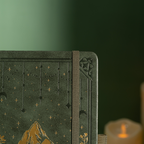 Close-up detail of the dark green vegan leather notebook cover, showcasing the embossed star and moon design, gold foil mountain peaks, intricate corner border, and a matching green elastic band, with a soft candle glow in the background. 