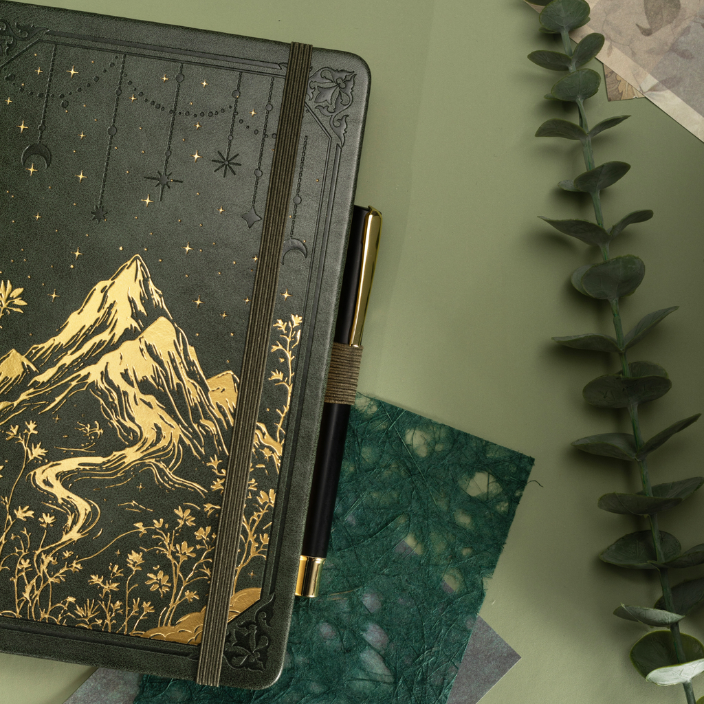 Forest green vegan leather journal with gold mountain embossing, styled in a nature-inspired setting with greenery and a pen. 