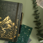 Forest green vegan leather journal with gold mountain embossing, styled in a nature-inspired setting with greenery and a pen. 