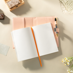 Open orange vegan leather notebook lying flat on a beige surface, showing the lined paper interior with an orange ribbon bookmark, a visible side pen loop holding a patterned pen, and a metal lock strap, styled with wooden stamps and fresh flowers.