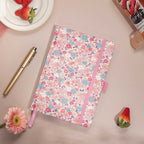 Flat lay of the pink ditsy floral notebook on a pink surface, featuring small blue and pink flowers and bright red strawberries.