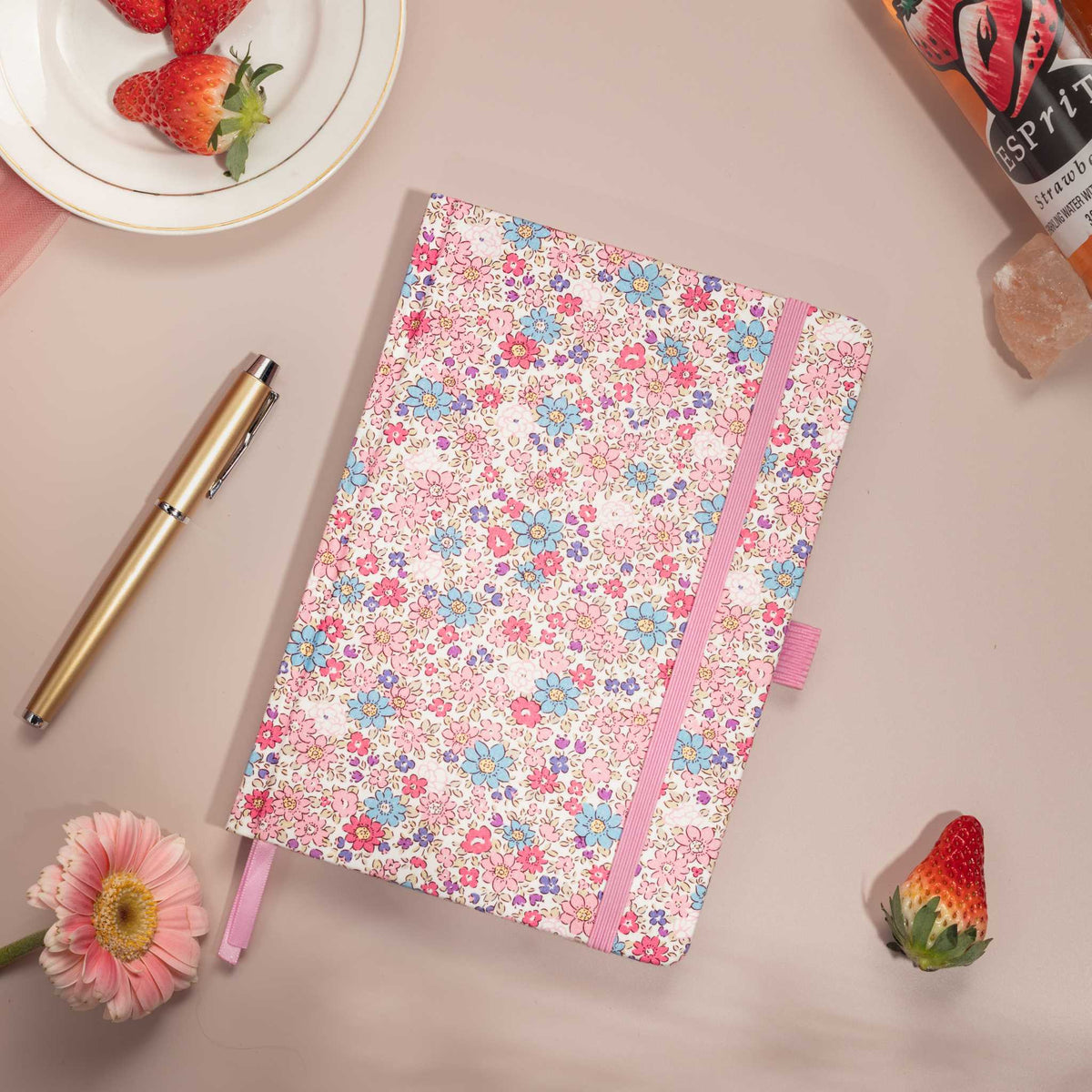 Flat lay of the pink ditsy floral notebook on a pink surface, featuring small blue and pink flowers and bright red strawberries.