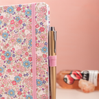 Side view of the pink ditsy floral fabric notebook standing upright, showing a pink and gold pen tucked into the side loop, styled with fresh strawberries on a pink desk.