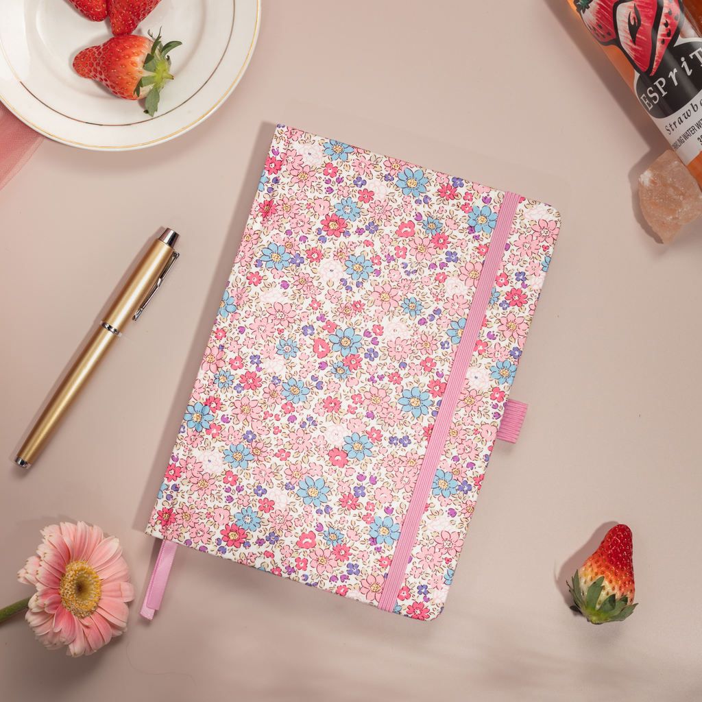 Flat lay of the pink ditsy floral notebook on a pink surface, featuring small blue and pink flowers and bright red strawberries.