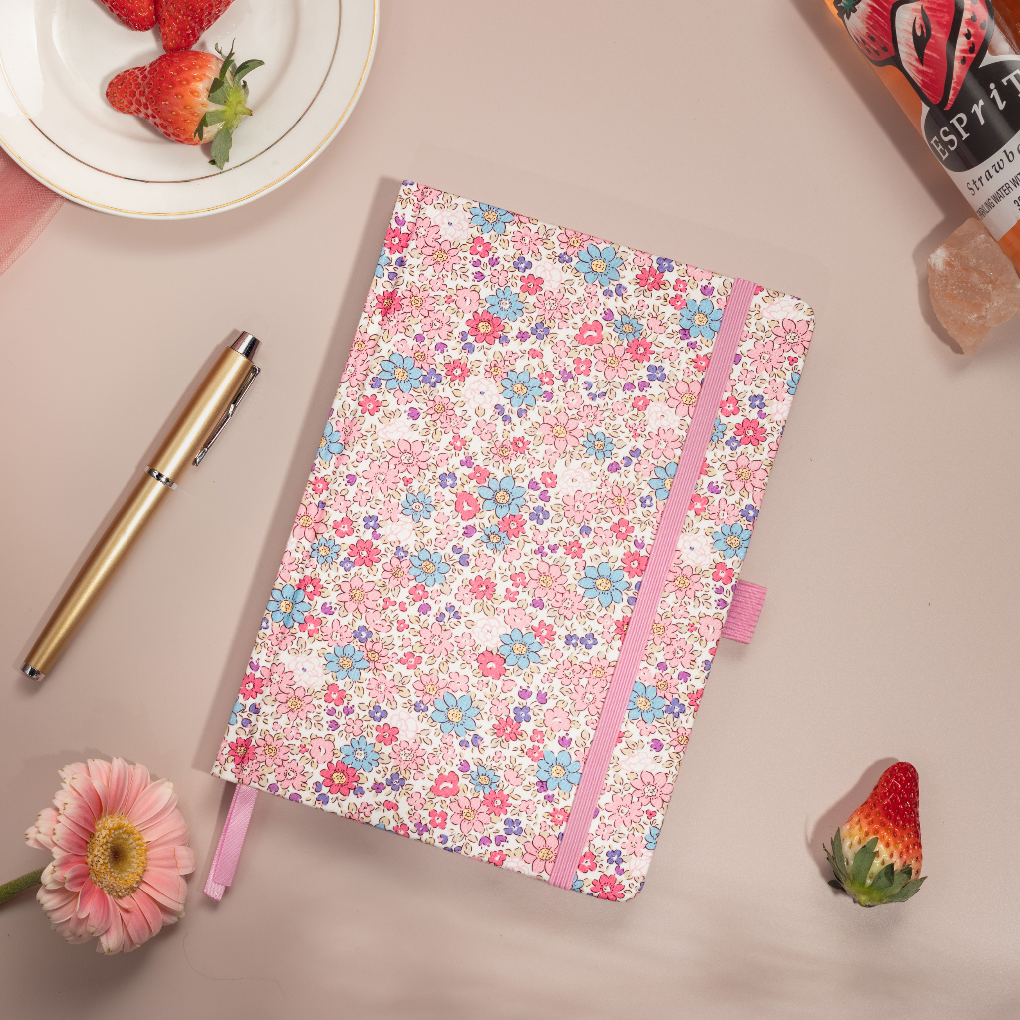 Flat lay of the pink ditsy floral notebook on a pink surface, featuring small blue and pink flowers and bright red strawberries.
