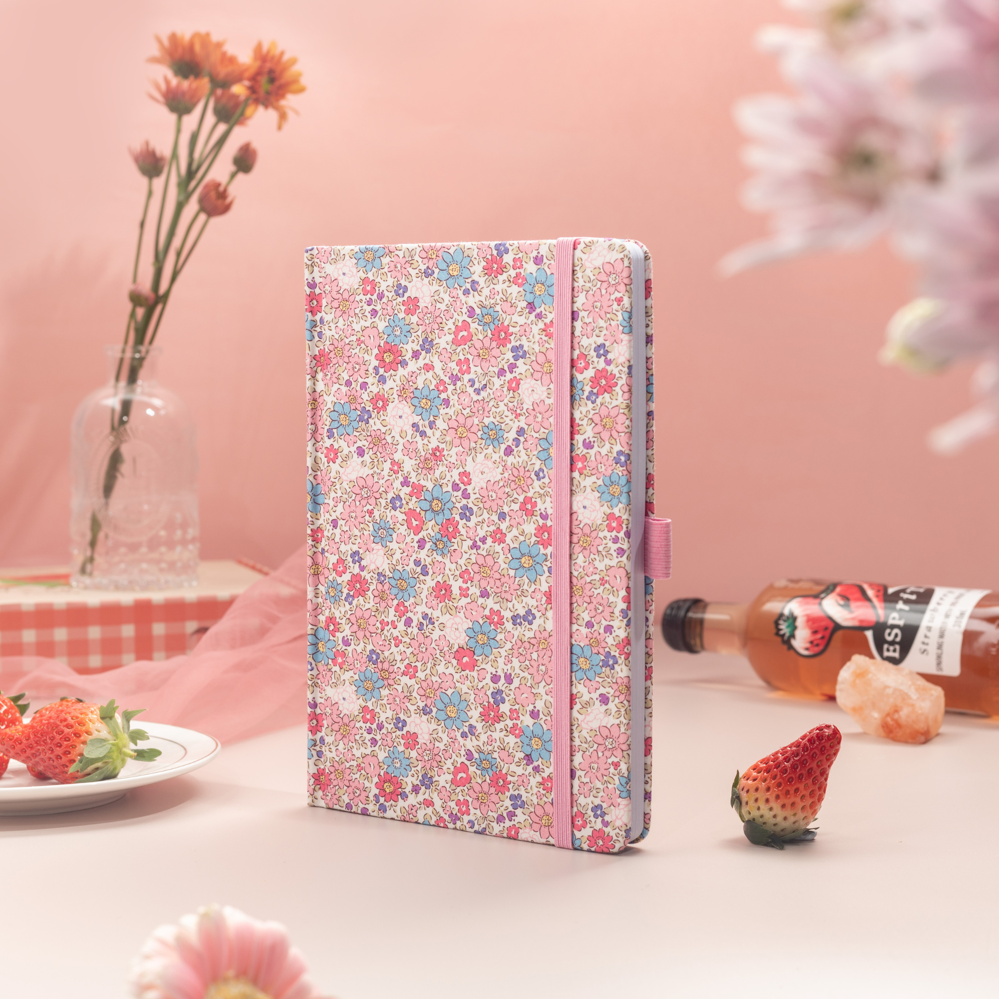 Pink ditsy floral fabric notebook standing upright on a pink desk, styled with fresh strawberries and a bottle of floral drink.