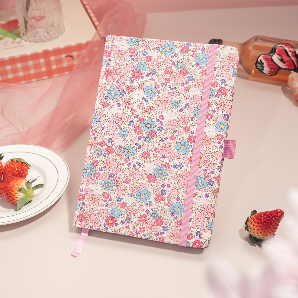Pink ditsy floral fabric notebook standing angled on a pink desk, with a matching pink elastic band and ribbon, styled with fresh strawberries and a strawberry drink bottle.