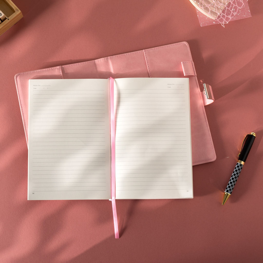 Open pink vegan leather journal lying flat on a pink surface, revealing the lined paper interior with Date and Memo No. headers, a pink ribbon bookmark, and a metal lock strap visible on the back cover, styled with a patterned pen.