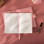 Open pink vegan leather journal lying flat on a pink surface, revealing the lined paper interior with Date and Memo No. headers, a pink ribbon bookmark, and a metal lock strap visible on the back cover, styled with a patterned pen.