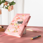 Pink floral vegan leather journal with a metal password lock, angled on a pink desk with a pen and flower vase.