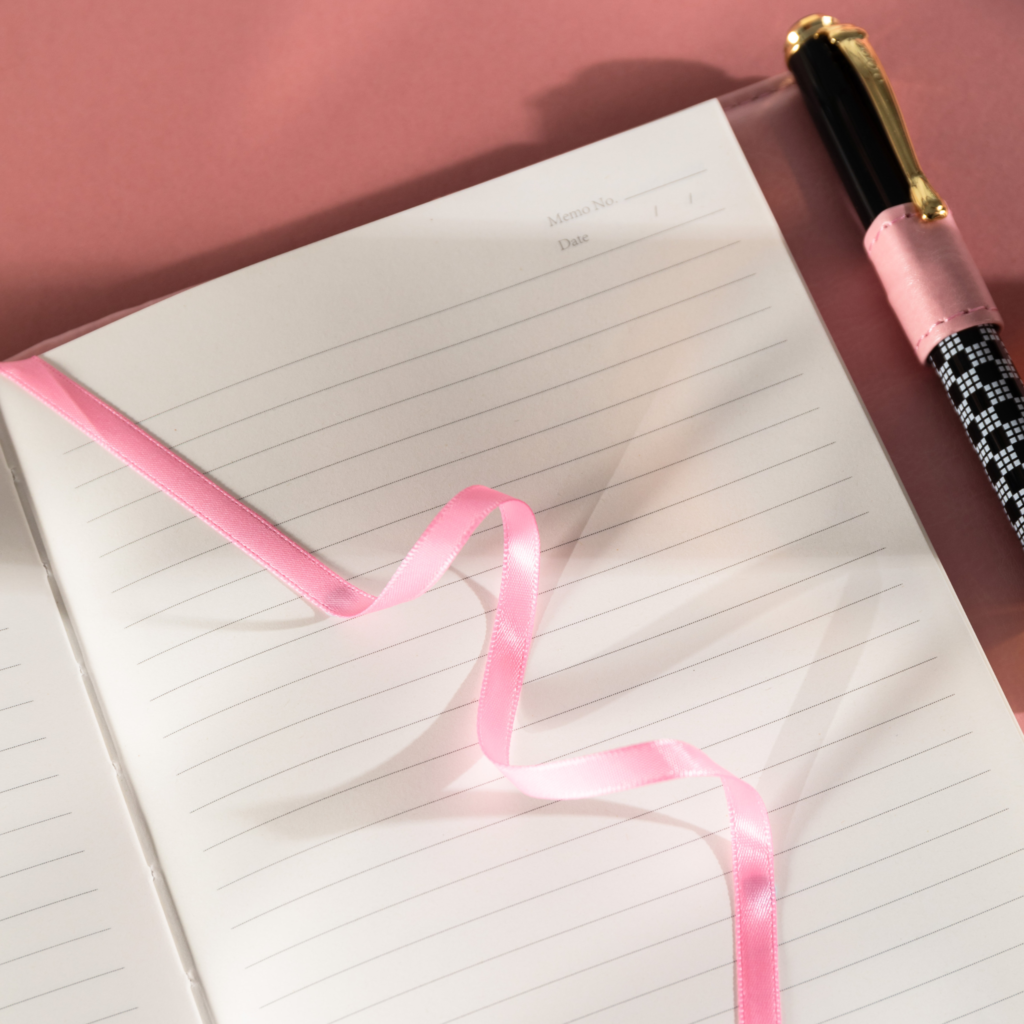 Close-up of the open lined paper interior with a pink satin ribbon bookmark resting on the page, next to a black and white pen. 
