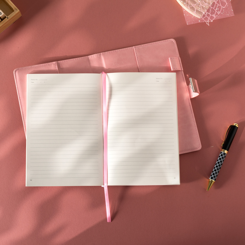 Open pink vegan leather notebook lying flat on a pink surface, revealing the lined paper interior with Date and Memo No. headers, a pink ribbon bookmark, and a metal lock strap visible on the back cover, styled with a patterned pen.