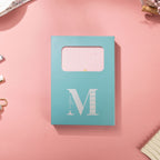 Light blue window-cut gift box featuring a white "M" logo, displaying the pink mountain vegan leather notebook inside.
