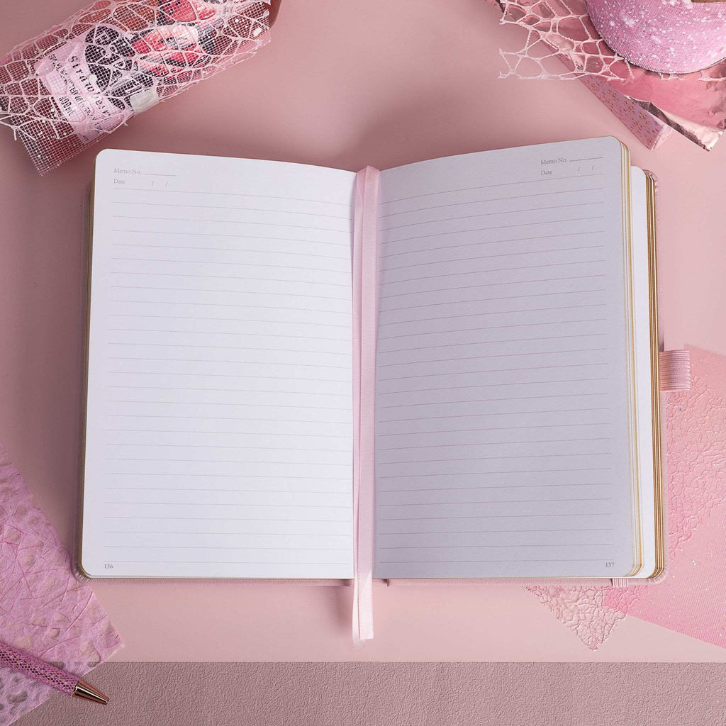 A5 line notebook opened flat on a pink desk, displaying clean white pages with a pink ribbon bookmark.