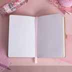 A5 line notebook opened flat on a pink desk, displaying clean white pages with a pink ribbon bookmark.