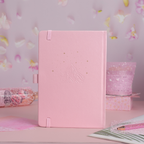 Back cover of the pink vegan leather notebook featuring elegant embossed gold foil stars, moon, and botanical details on a pink desk setup.