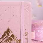 Close-up of the pink vegan leather notebook cover showing gold foil mountain and starry night embossing with a matching pink elastic band.