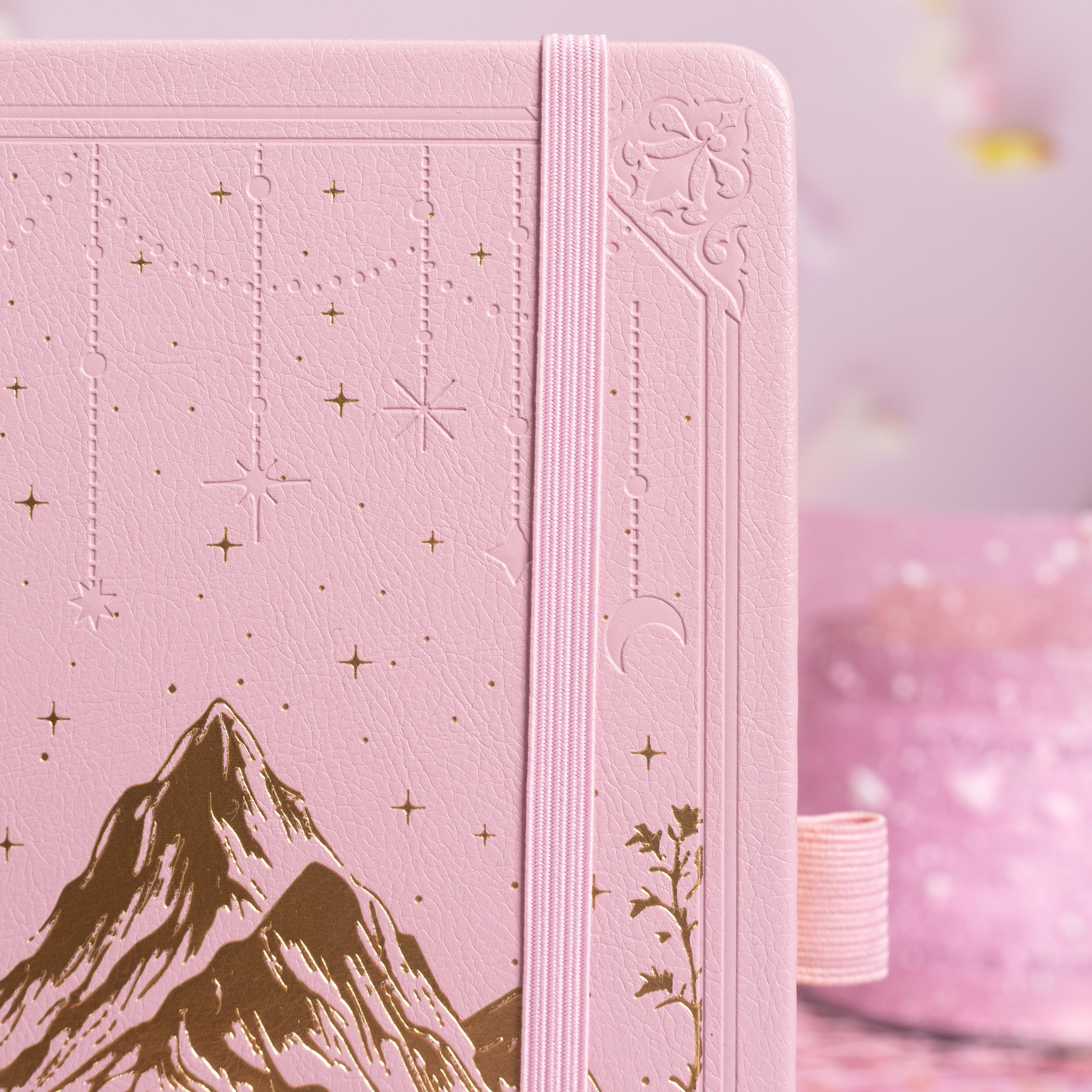 Close-up of the pink vegan leather notebook cover showing gold foil mountain and starry night embossing with a matching pink elastic band.