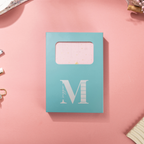 Light blue window-cut gift box featuring a white "M" logo, displaying the pink mountain vegan leather notebook inside.