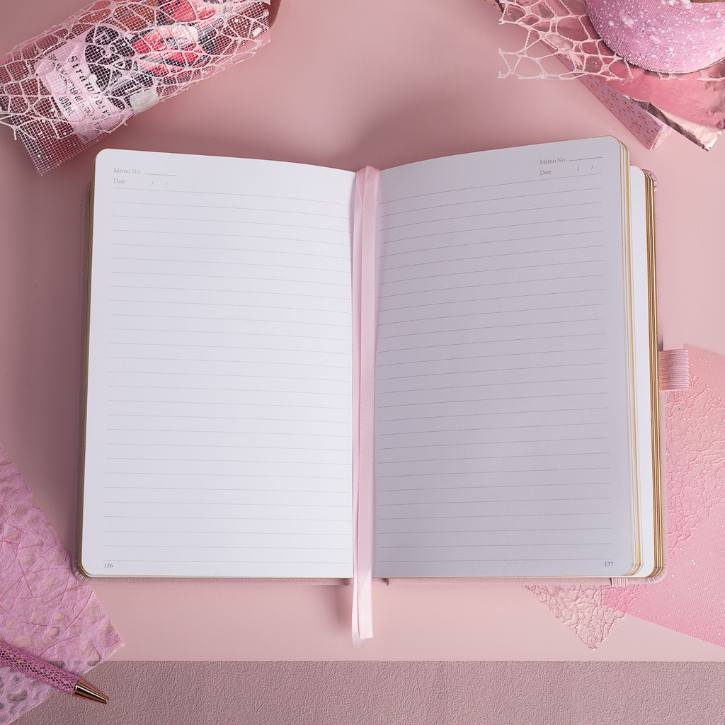 A5 line notebook opened flat on a pink desk, displaying clean white pages with a pink ribbon bookmark.