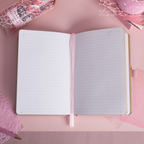 A5 line notebook opened flat on a pink desk, displaying clean white pages with a pink ribbon bookmark.