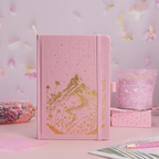 Pink vegan leather journal with a gold foil mountain scene standing upright on a pink desk with a pen and floral gift boxes.