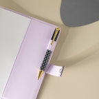 Close-up of the purple vegan leather journal, showing a black and white patterned pen tucked into the side pen loop, with the metal lock strap extended and visible stitching details on the cover.