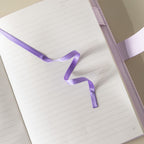 Close-up of the open lined paper interior with a purple satin ribbon bookmark and a pen.