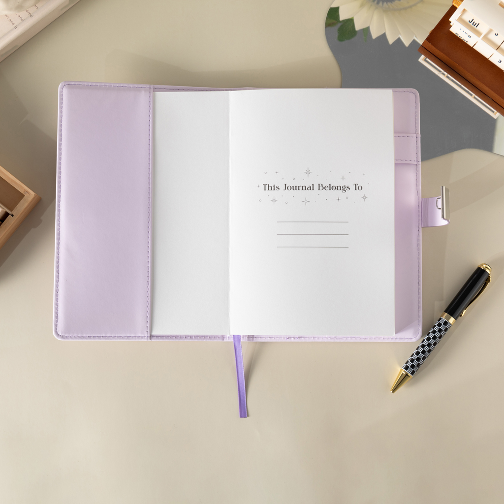 Open purple vegan leather notebook showing the "This Journal Belongs To" ownership page, laid flat with a matching purple ribbon and a pen.