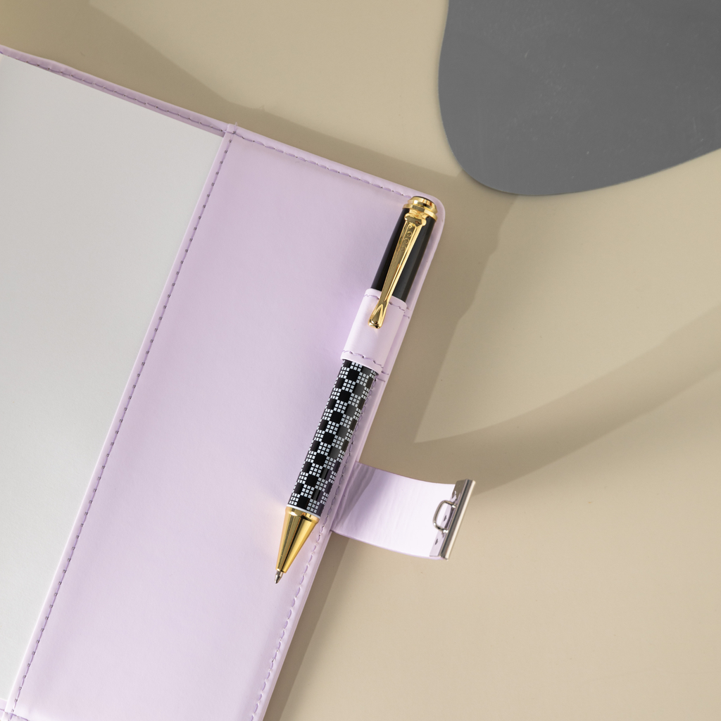 Close-up of the purple vegan leather notebook, showing a black and white patterned pen tucked into the side pen loop, with the metal lock strap extended and visible stitching details on the cover.