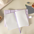 Open purple vegan leather notebook lying flat on a beige surface, showing the lined paper interior with a purple ribbon bookmark, an open metal lock strap, and styled with a book and a floral fan.