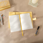 Top-down view of the open yellow vegan leather journal lying flat on a beige desk, featuring lined paper with a yellow ribbon bookmark, an open metal lock strap, and styled with a patterned pen and a spiral journal.