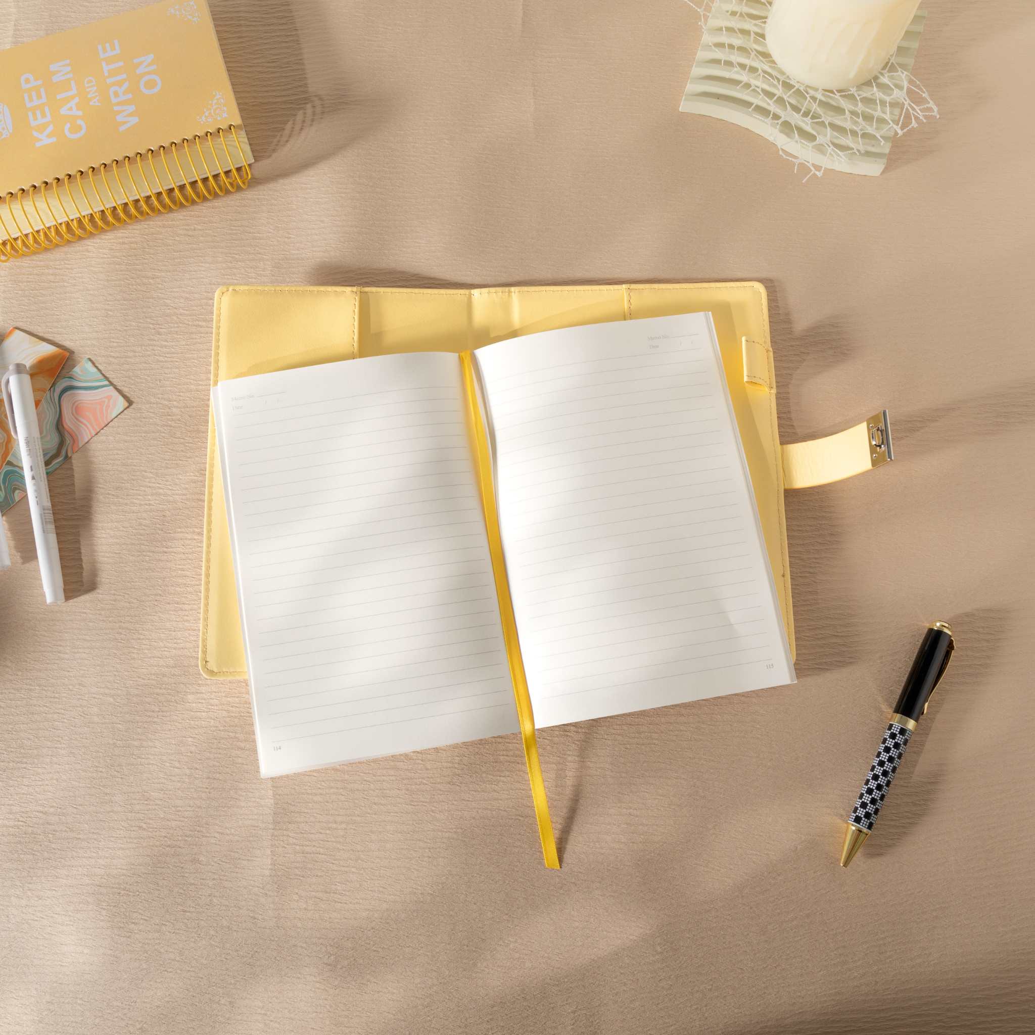 Top-down view of the open yellow vegan leather journal lying flat on a beige desk, featuring lined paper with a yellow ribbon bookmark, an open metal lock strap, and styled with a patterned pen and a spiral journal.