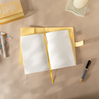 Top-down view of the open yellow vegan leather notebook lying flat on a beige desk, featuring lined paper with a yellow ribbon bookmark, an open metal lock strap, and styled with a patterned pen and a spiral notebook.